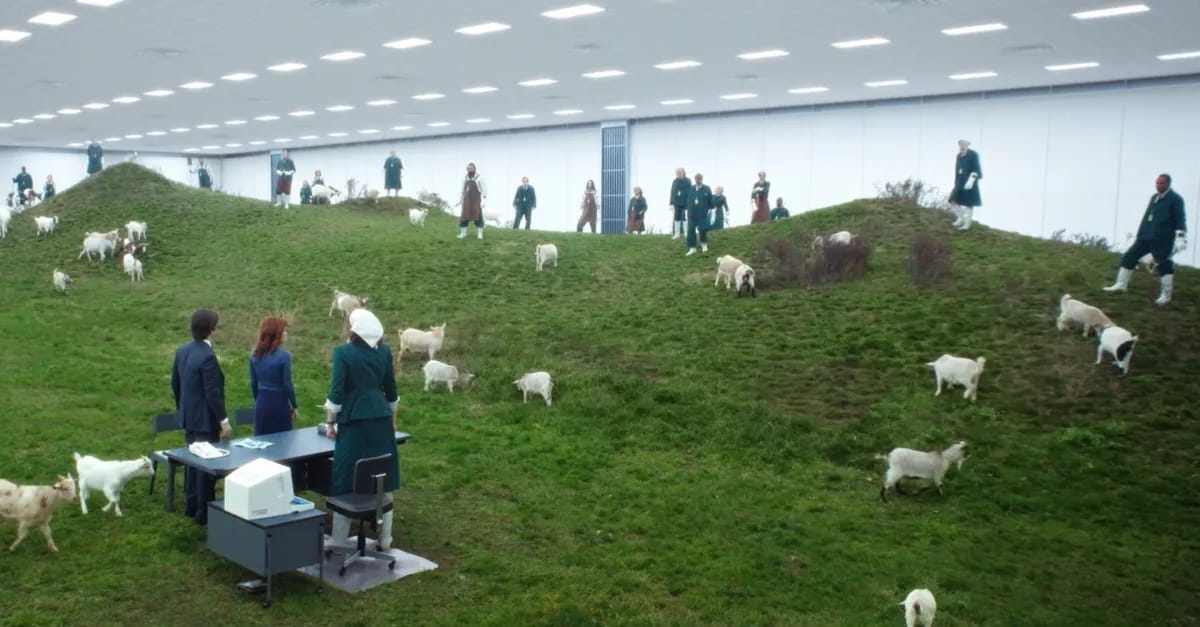 Still from Severance. People stood along the crest of a hill, goats everywhere, looking down at 3 people stood by a desk